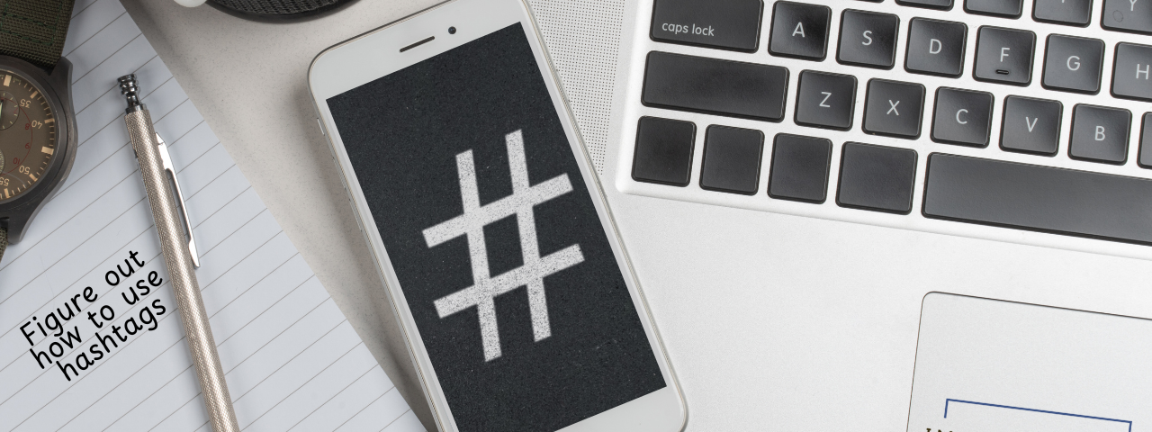 Hashtag symbol on a phone with a laptop and a piece of paper that says figure out how to use hashtags