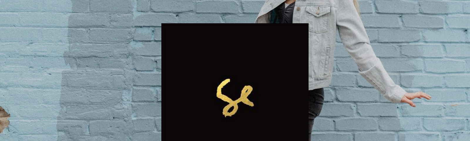 blonde woman walking making big steps ahead of a light blue brick wall, in the foreground is the album cover of Sylvan Esso, by duo Sylvan Esso