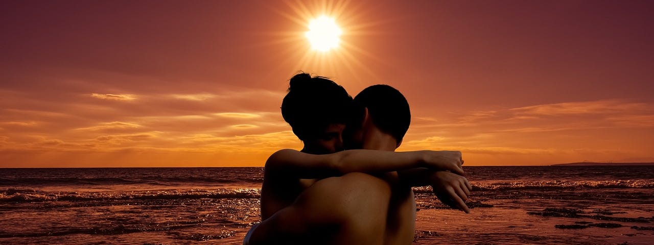A pair of naked lovers standing in each others arms at the waters edge on the beach as the sun sets in the background