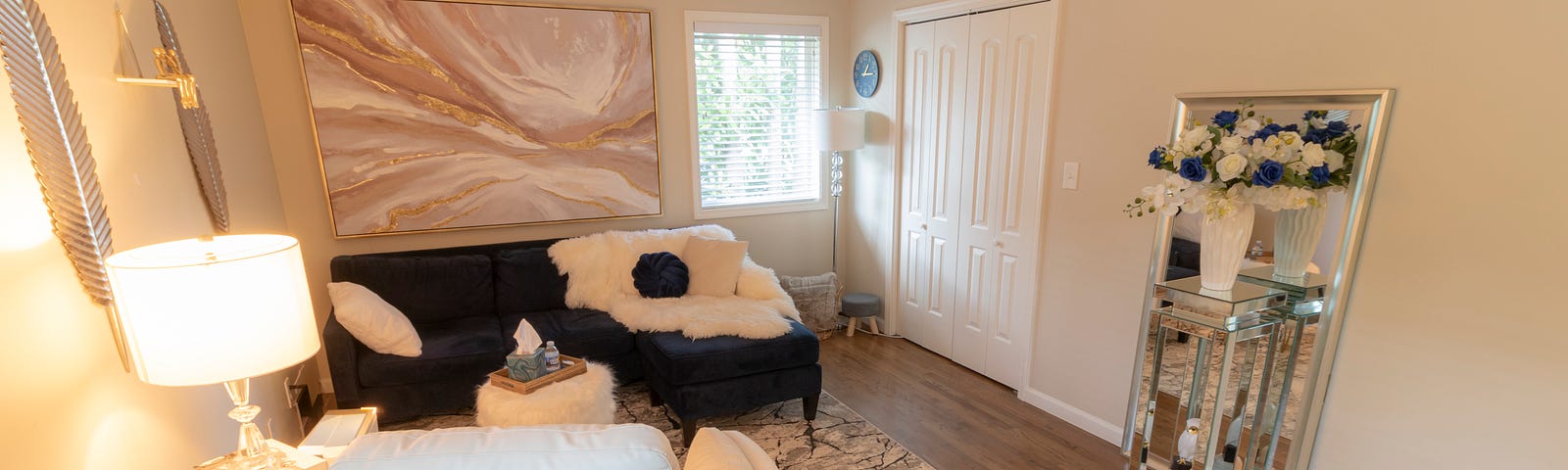 A Cozy therapy office with a blue couch and soft blanket. A white chair is in the foreground and the back wall in has an abstract painting. The room is a warm amber blow