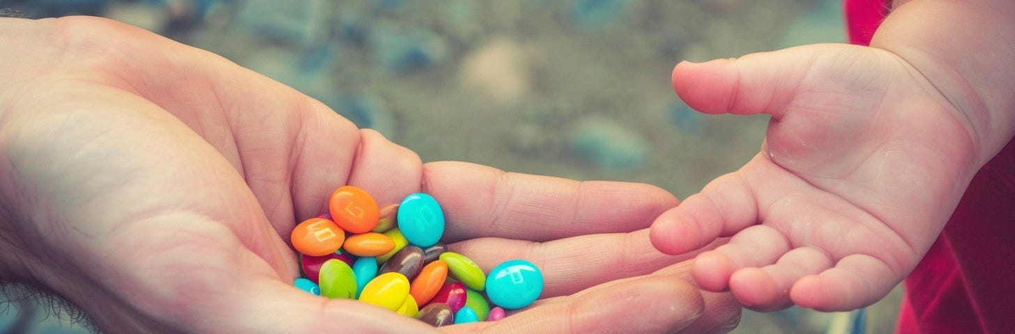 Two hands, one belonging to an adult and one to a child, holding bright, multicolored candies warmly shared between them.