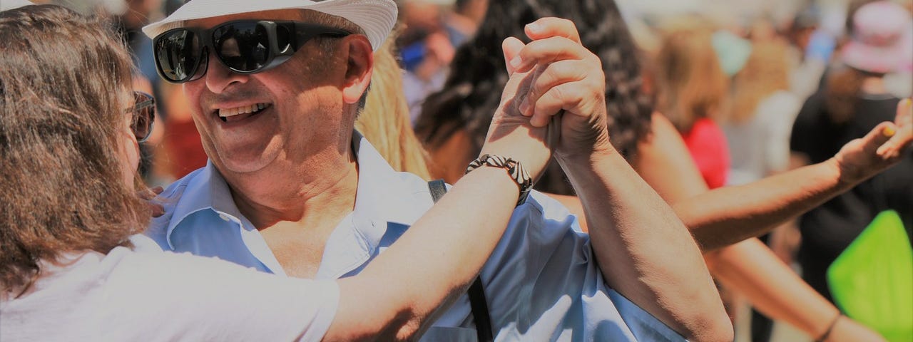 A man in his sixties wearing a white hat, sun glasses, and blue shirt dances with a woman in a white blouse.