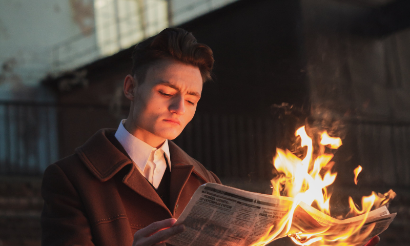 A young man in a leather coat is calm, reading a newspaper. The newspaper is on fire.
