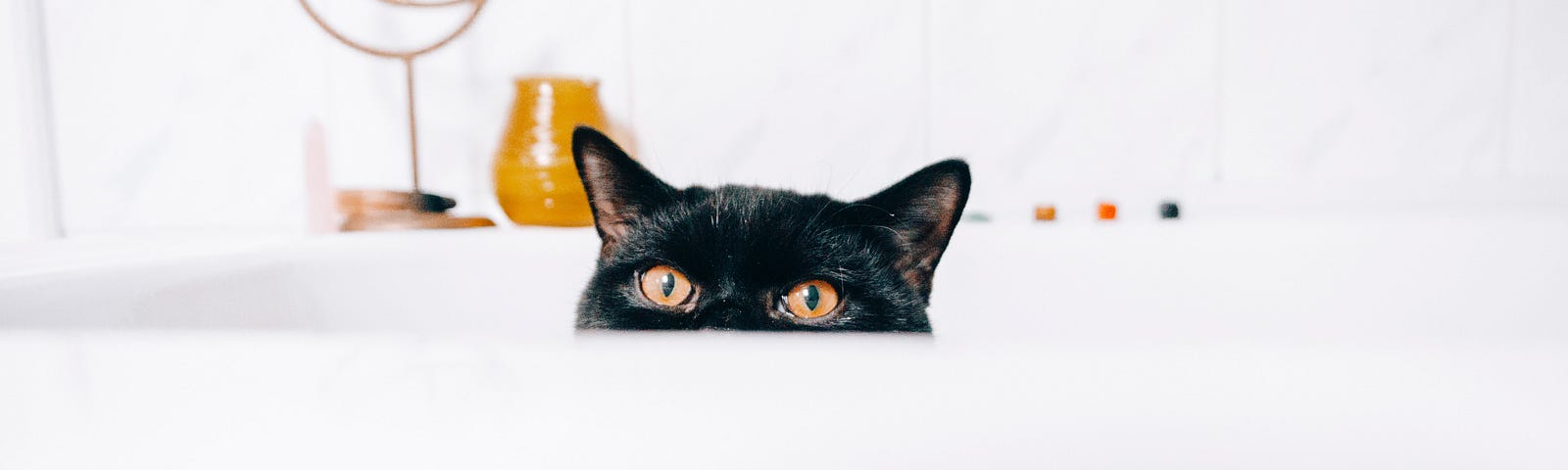 A black cat peeking out of a white bathtub, only showing the top half of his face/head.