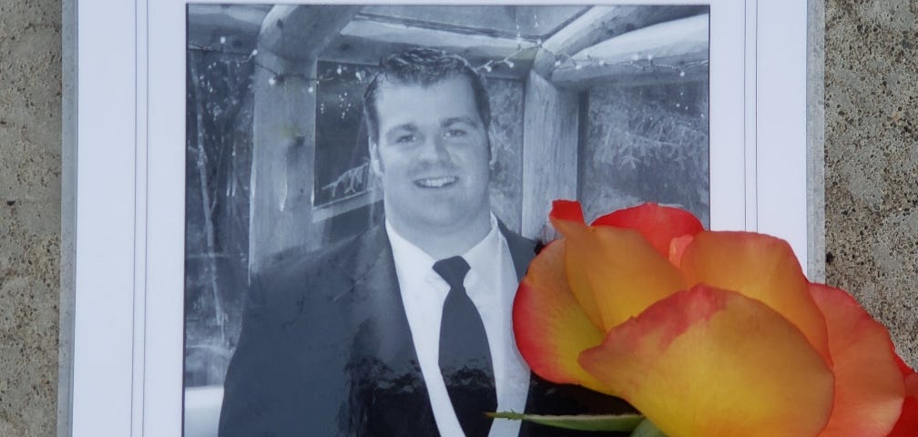 Black and white photo of a memorial brochure, showing Alex,  who died by suicide in 2012. With a yellow rose from the author’s garden…