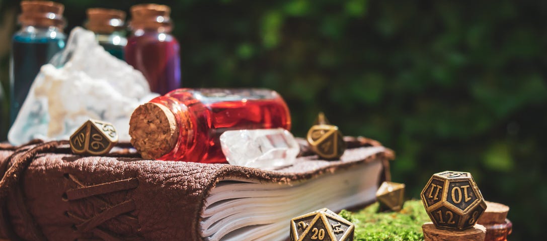 Image: A photo of a leather-bound journal surrounded by bronze dice, a large quartz crystal, and several corked potion bottles.