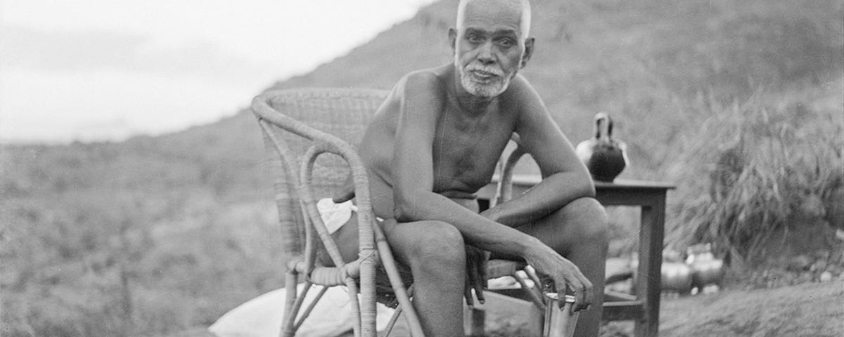 Indian mystic Ramana Maharshi (1879–1950) sits in a chair bending over, with the holy hill Arunachala in the background. Image in black & white.