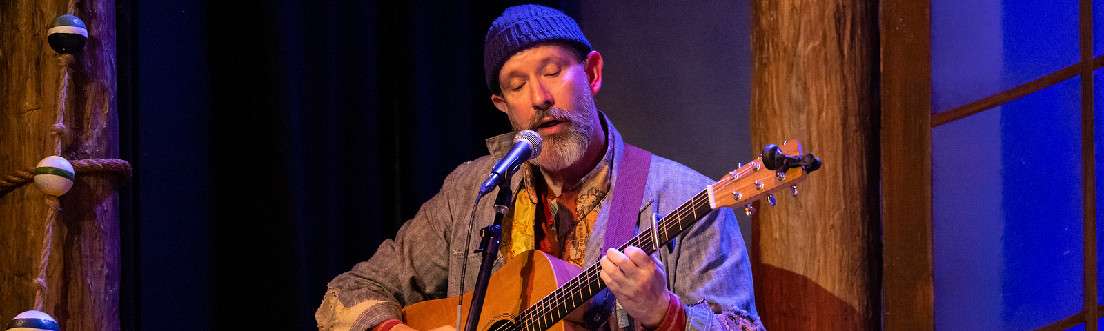 A performer with a beard, in a beanie and denim coat, plays guitar and sings into a microphone.