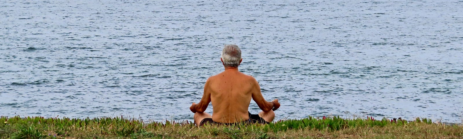 Older man sits on the grass in the lotus position, meditating as he looks out to sea.