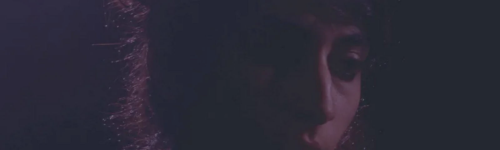 A screenshot of Arooj Aftab, a Pakistan-born, Brooklyn-based composer and singer. This image is a quarter profile photograph where she is back lit and bathed in dark purplish tones that resembles a chiaroscuro painting.