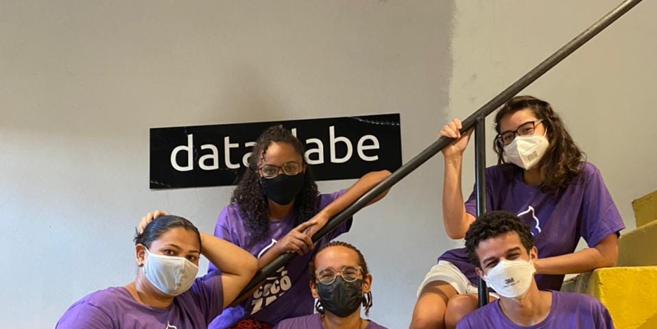 Embaixadores Cocôzap. Na foto estão 5 jovens, sendo 3 mulheres e 2 homens. Todos estão vestidos com uma camiseta roxa escrito "Cocôzap" e usam máscaras de proteção por conta da COVID-19. Estão posando em frente a uma escada amarela, e no fundo, há uma placa na parede escrito "data_labe".