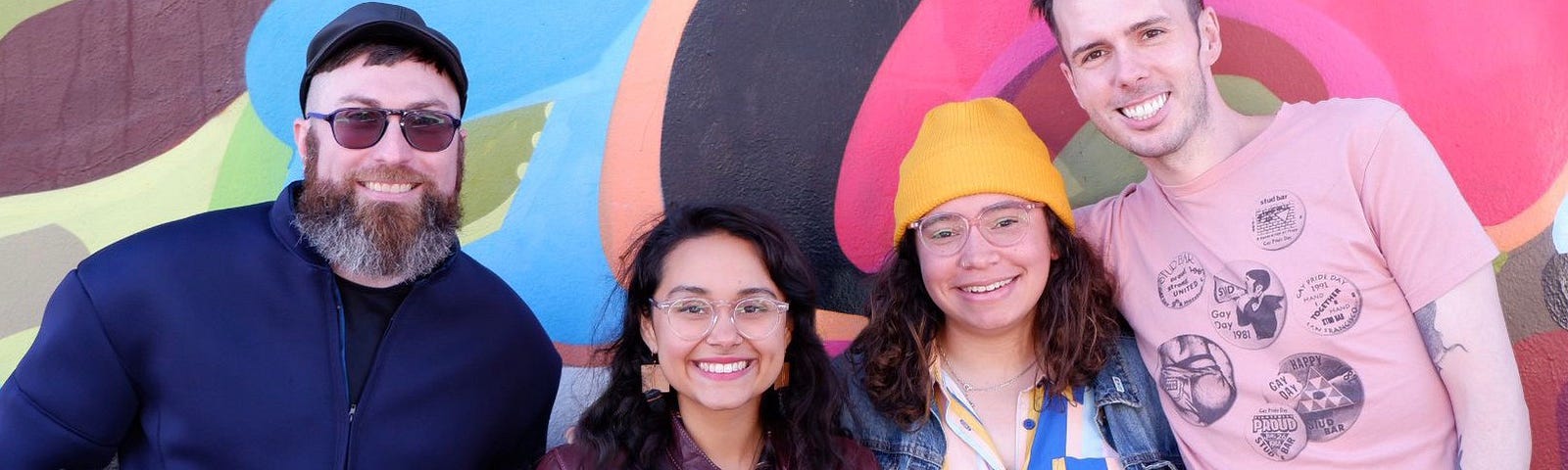 A smiling group of four queer design club members in front of a colorful mural in the Mission.
