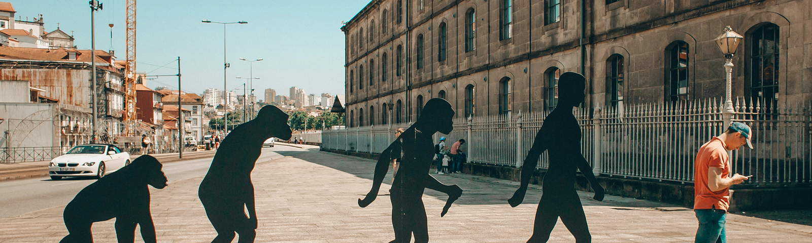 An art installation of four black metal silhouettes depicting hominid evolution, progressing from left to right, and a man dressed in blue jeans, a red t-shirt and a cap in the last position, looking down at his phone in his hand. The installation is in Porto, Portugal, on a sunny day, with a clear blue sky and tower cranes, apartment buildings, street lights and a car in the background to the left, and what looks to be a large old brick warehouse in the background on the right.