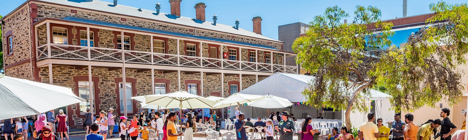 Crowd of people in a courtyard with marquee and umbrellas set up in front of a two story bluestone building.