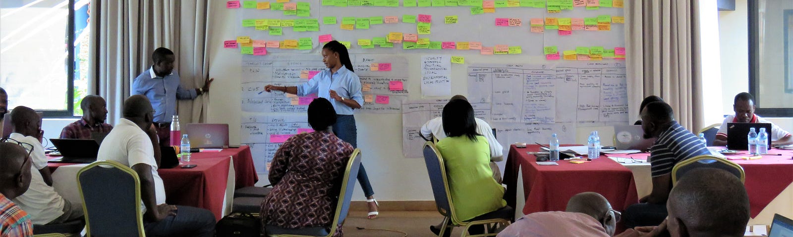 Two people stand in front of groups of people sitting at tables at a workshop. The two people in the front are pointing to a wall with paper and post-it notes taped to it.
