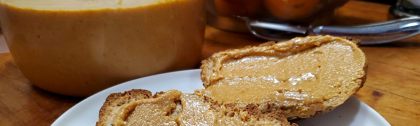 White plate with two slices of bread with peanut butter sitting on a wooden surface in front of a plastic container with peanut butter in front of a wire basket of oranges and a vegetable peeler.