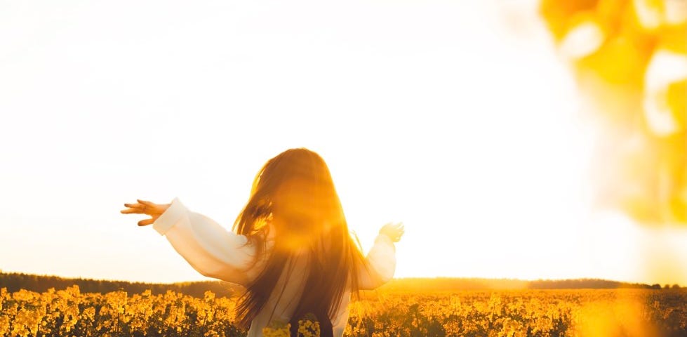 A woman with long dark hair runs through a field of yellow flowers as the sun rises.
