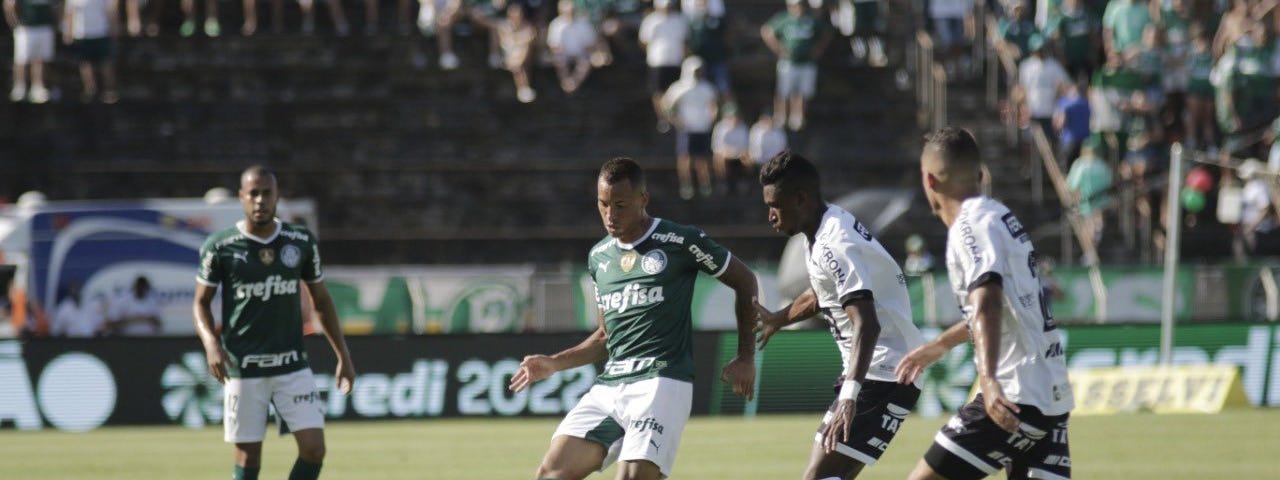 Cena do jogo Palmeiras x Inter de Limeira, pelo Paulistão; Breno Lopes, jogador do Palmeiras, de camisa e meias verdes e calção branco, chuta de é direito observado de perto por dois jogadores da Inter, de camisa e meias brancas e calções pretos. Jogo de ida, sob sol forte.