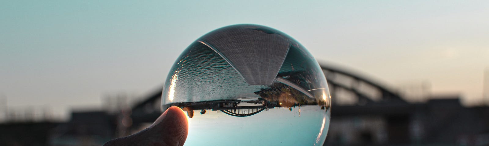 A hand holds up a glass sphere, showing an upside down perspective of a bridge over water.