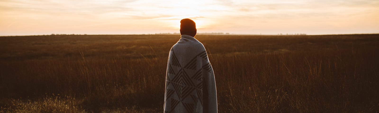 A person, wrapped in a blanket, standing in a field at golden hour.