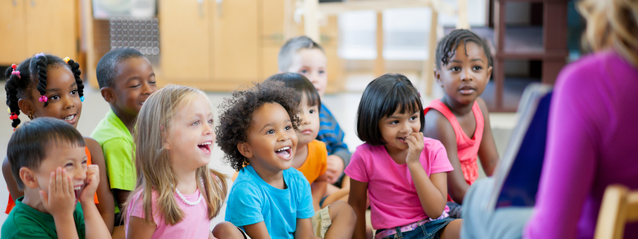 Kindergarteners sit on the floor in their classroom listening to their teacher reading a story.