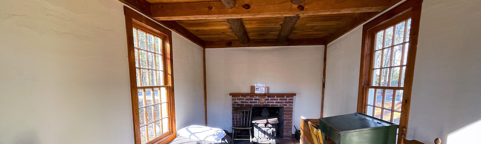 A single room cabin with a table and bed on the left, writing desk and chair on the right, and wood stove with fire place at the back wall. A window on either side. The cabin is built in an 1800s style with simple, old-fashioned furniture and wood frame, ceiling, and floors.