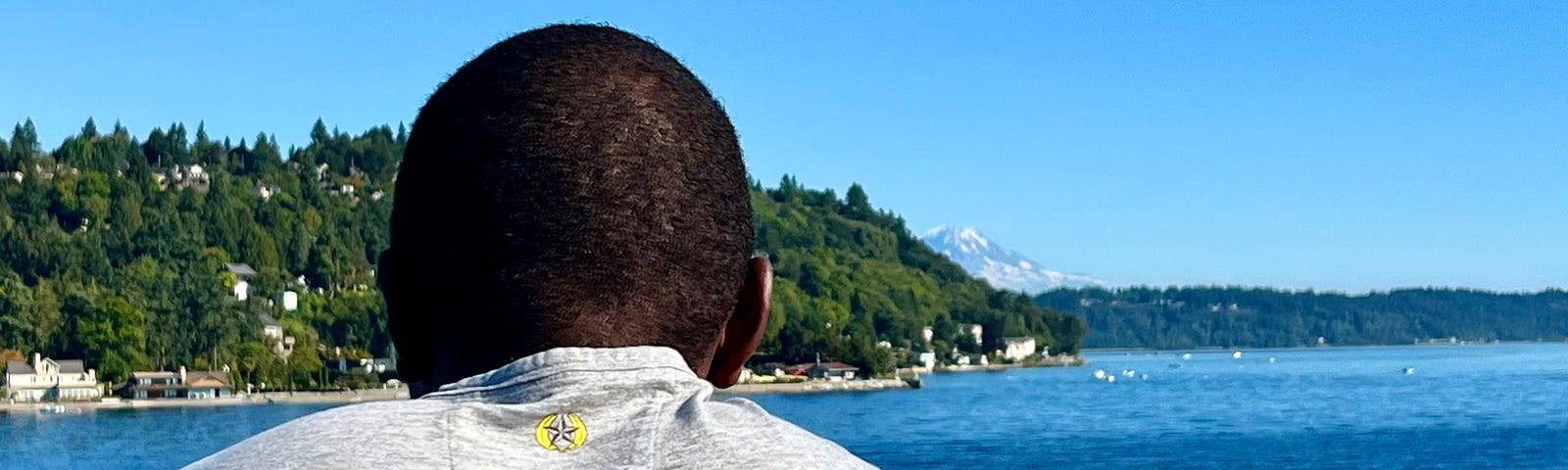 Back of a black man looking out over water with mountain and land in front of him.