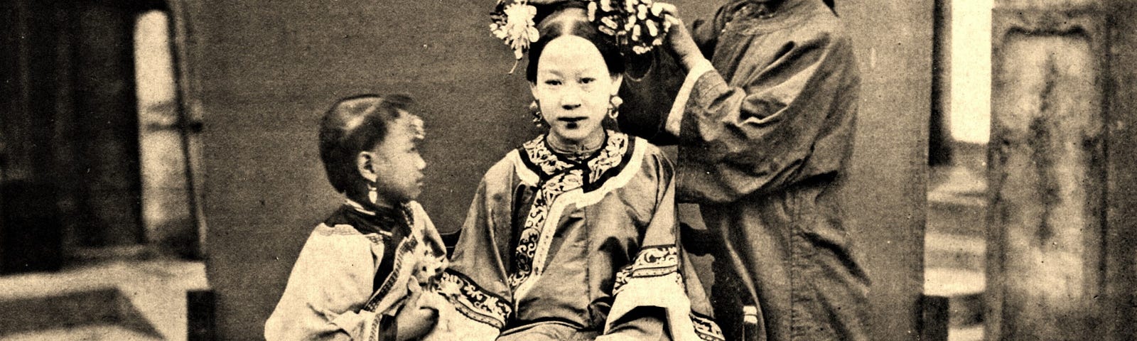 A lady sits still while a girl dresses her hair and a child tries to get her attention.