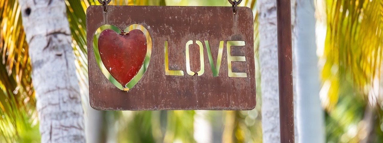 A metal sign with a red heart and cut out letters L O V E in front of palm trees.