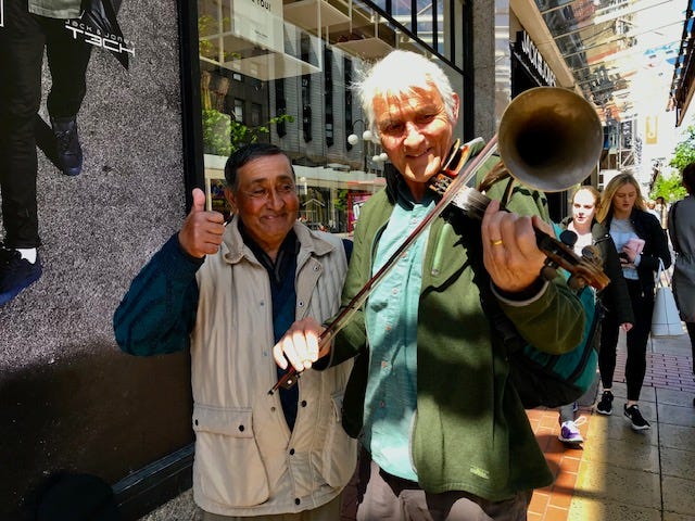 Two men standing on the sidewalk in downtown Belfast. Author’s husband is playing a street musician’s homemade string instrument similar to a violin