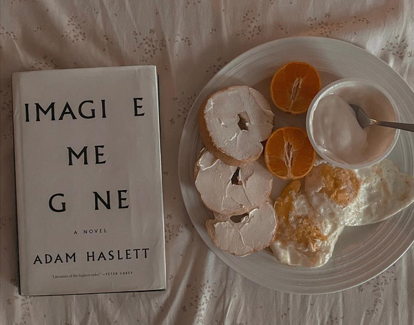 Flatlay photo of the hardcover copy of Imagine Me Gone by Adam Haslett. Next to the book is a plate of breakfast. Background is a white floral sheet.