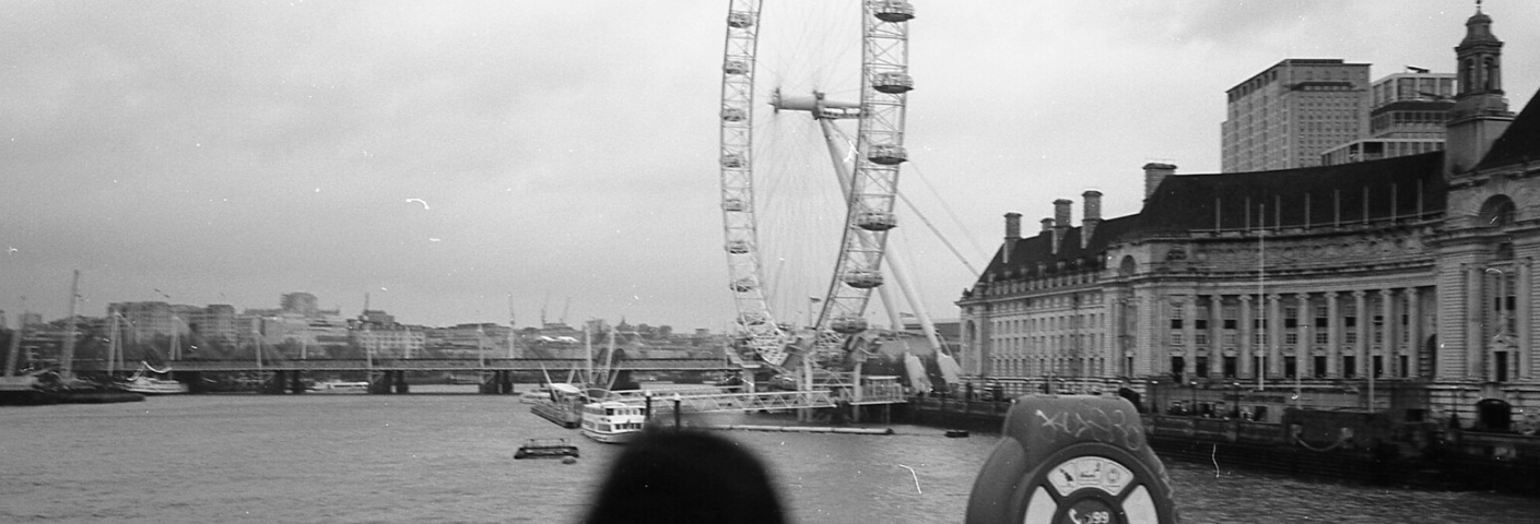 women looking out on the water from a bridge in London. You can see a Ferris wheel and different buildings. The picture is black and white