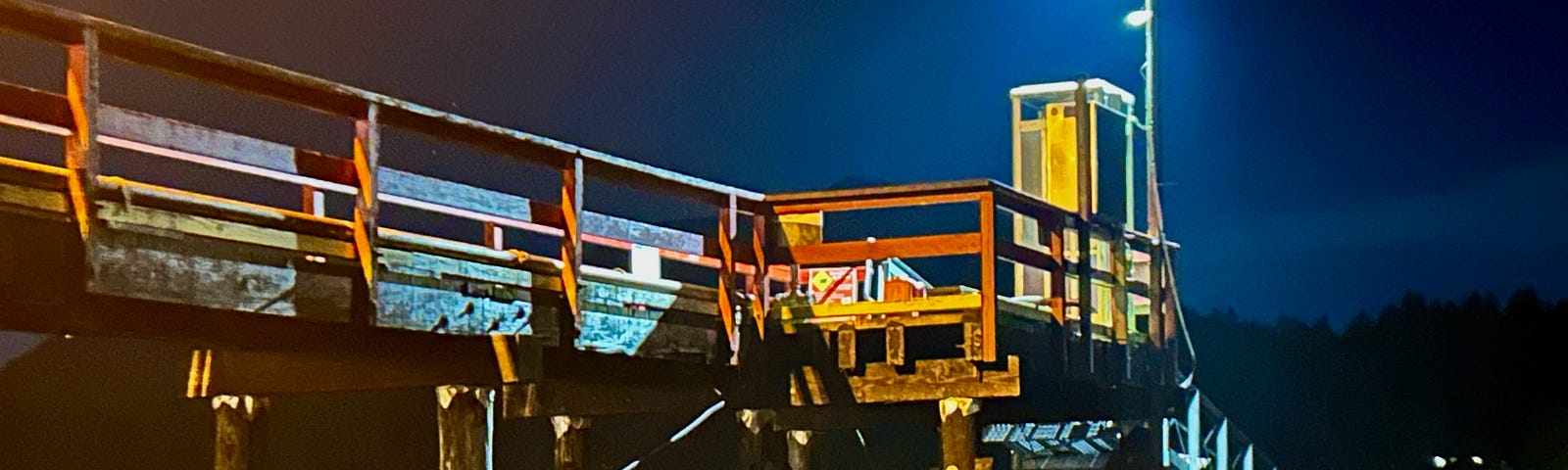 It is dark out. A walkway leads to a phone booth. Leading down from the phone booth is a sloping walkway that leads to a long dock. A light shines over the phone booth shining in a dark blue sky.