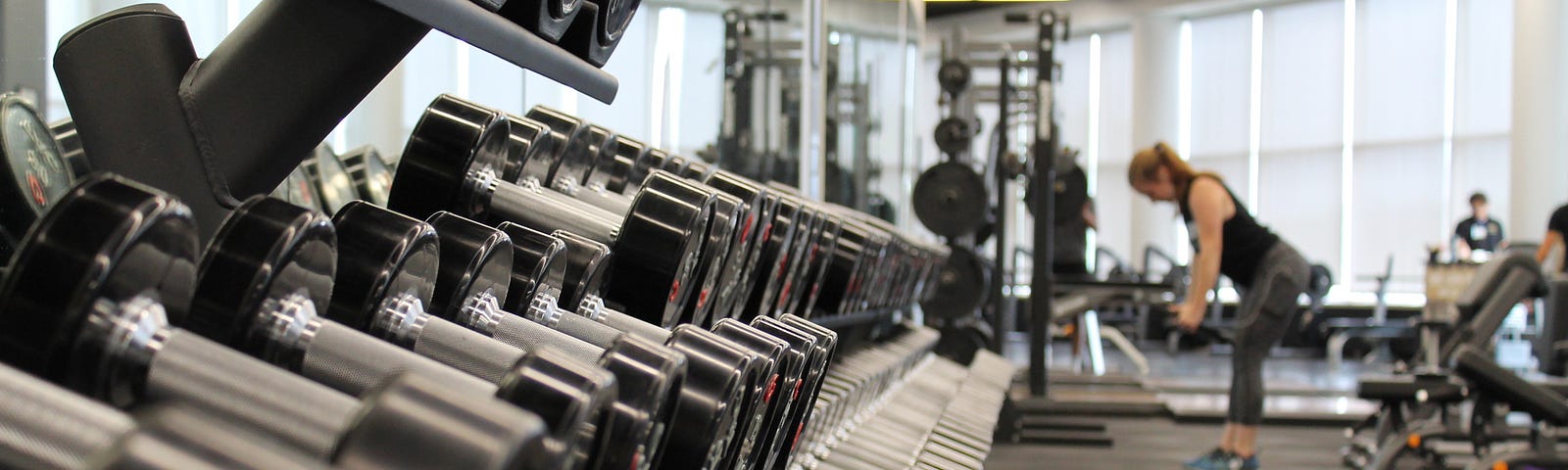 A photograph of a gym. In the foreground is a closeup of a rack of barbells, and in the background is a woman lifting weights.