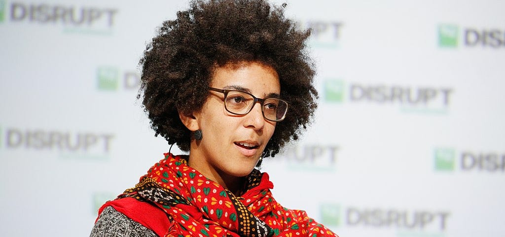 a person with glasses wearing a short sleeve top and a long wrap around scarf gesturing and talking while seated. In the background, there is a surface with the word “disrupt” written in a pattern