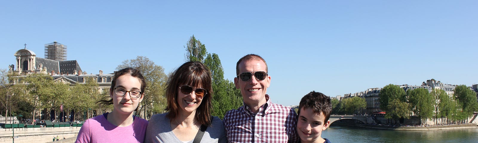 A family of four poses on a bridge over a river on a sunny day