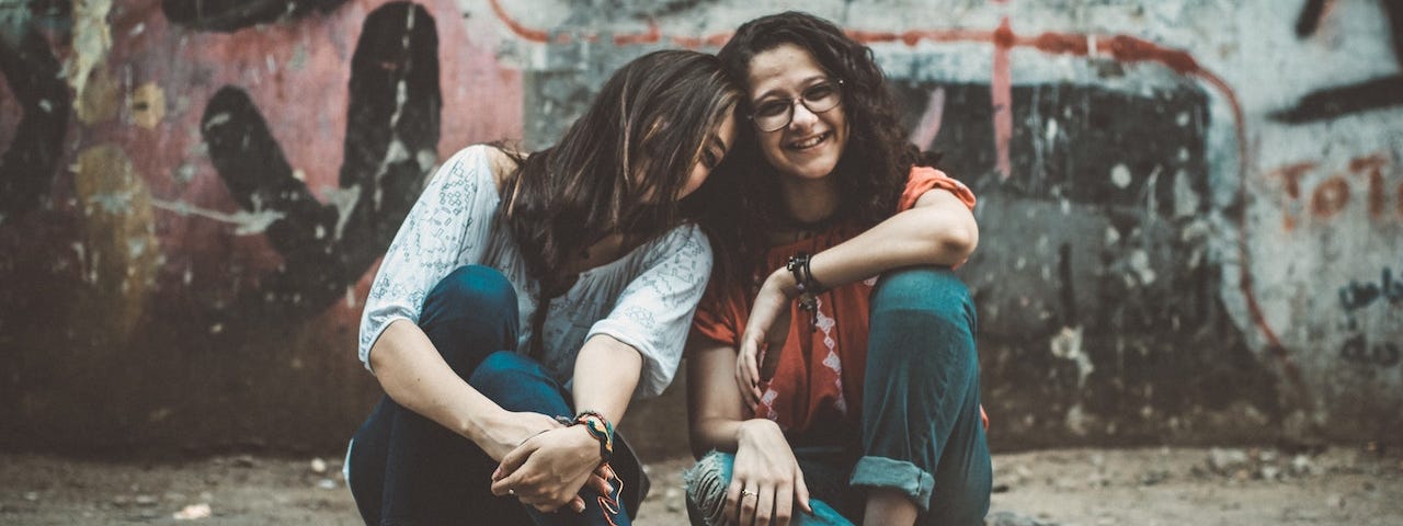 Two women sitting close together on a curb.