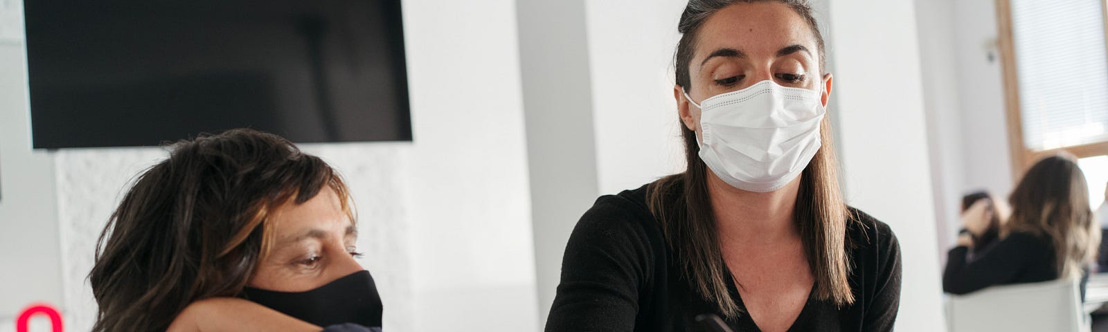 Dos mujeres con mascarilla sentadas en una mesa en un aula, con una pantalla de televisión detrás y cuadernos sobre la mesa.