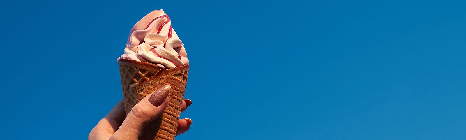 Hand holding up an ice cream cone. The background is a brilliant blue sky.