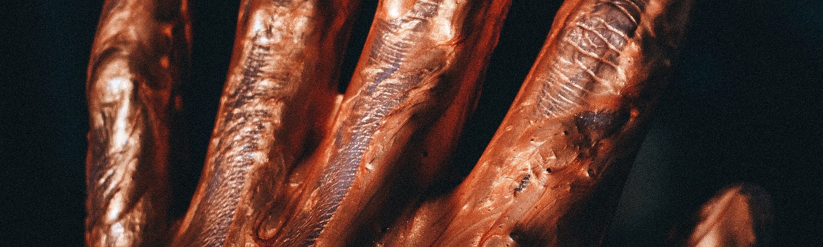 Dark close up of a human hand, covered in metallic orange paint with gooey strands stretching between each finger.