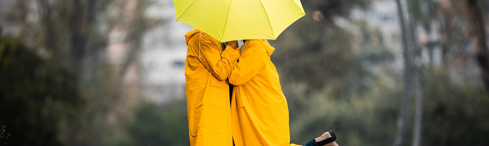 A couple stand facing each other. They are both wearing large, shapeless raincoats and a large umbrella obscures their faces, but from their poses we can guess they are kissing.