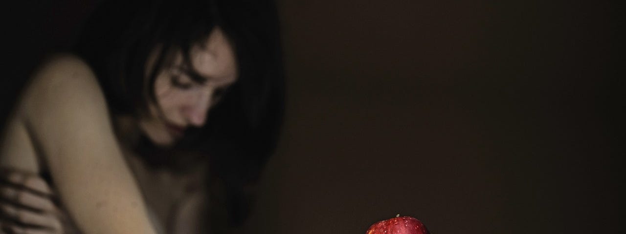 A person in shadow, head bowed, long dark hair falling over their face, one hand stretched out into the light with a red apple resting on their upturned palm.