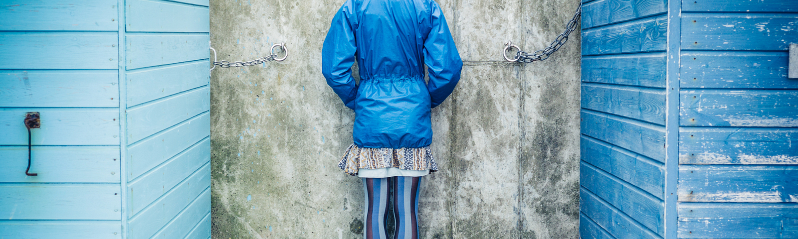 A person wearing a blue windbreaker with hands in pockets has their back turned to us and stands against a concrete wall. Two blue wooden structures are on either side of them.