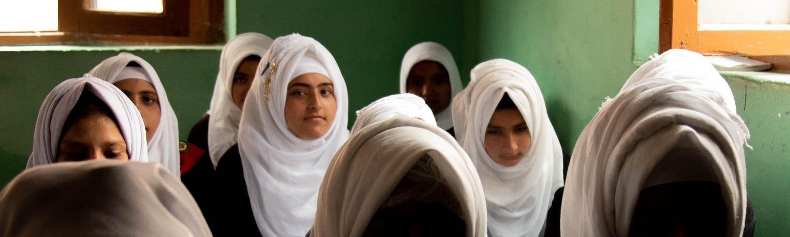Three rows of young women in white head scarves sit in a green room. Many of the women are looking down, while a few look directly at the camera.