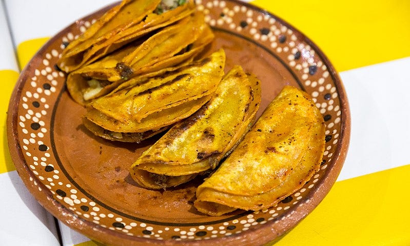 A brown ceramic plate with five crispy tacos on a yellow and white striped tablecloth.