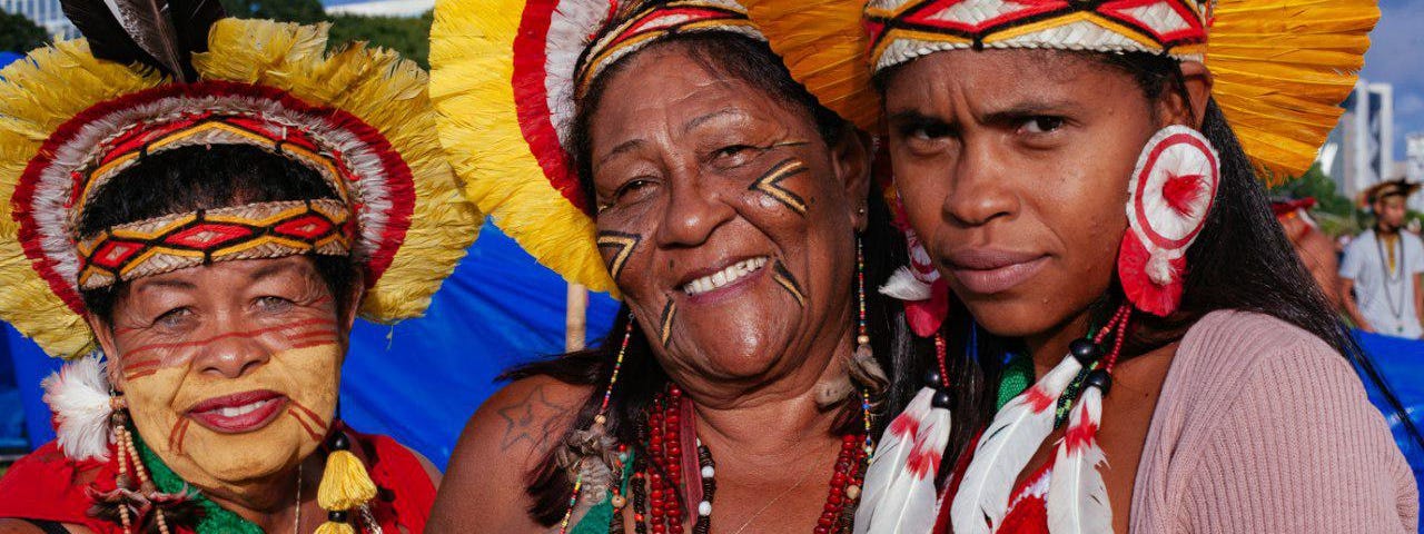 Três mulheres indígenas olha para a lente da câmera. Duas são mais velhas e sorriem. A mais jovem parece desconfiada