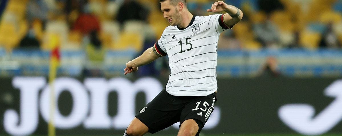 O jogador Niklas Süle chuta a bola, e está vestindo o uniforme da seleção alemã