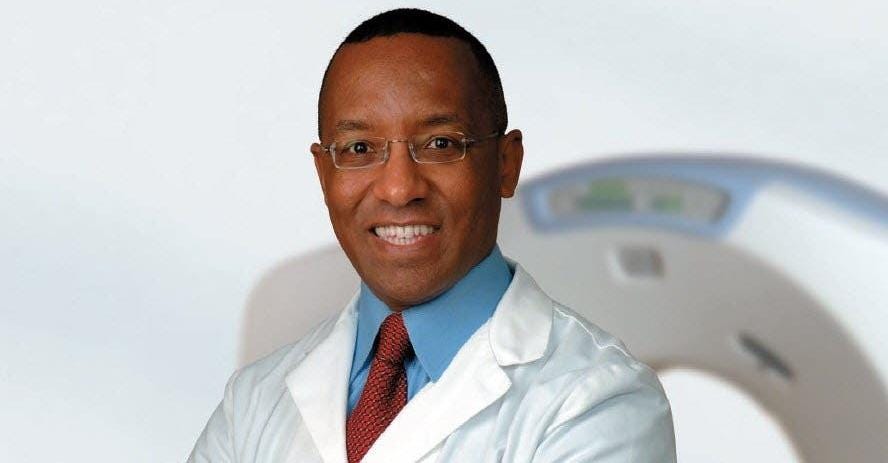 Photograph of the author, Dr. Michael Hunter, in a white doctor’s coat. He wears a blue shirt with a red tie and rimless eyeglasses. In the background is a CT scanner. In the far background is a white wall.