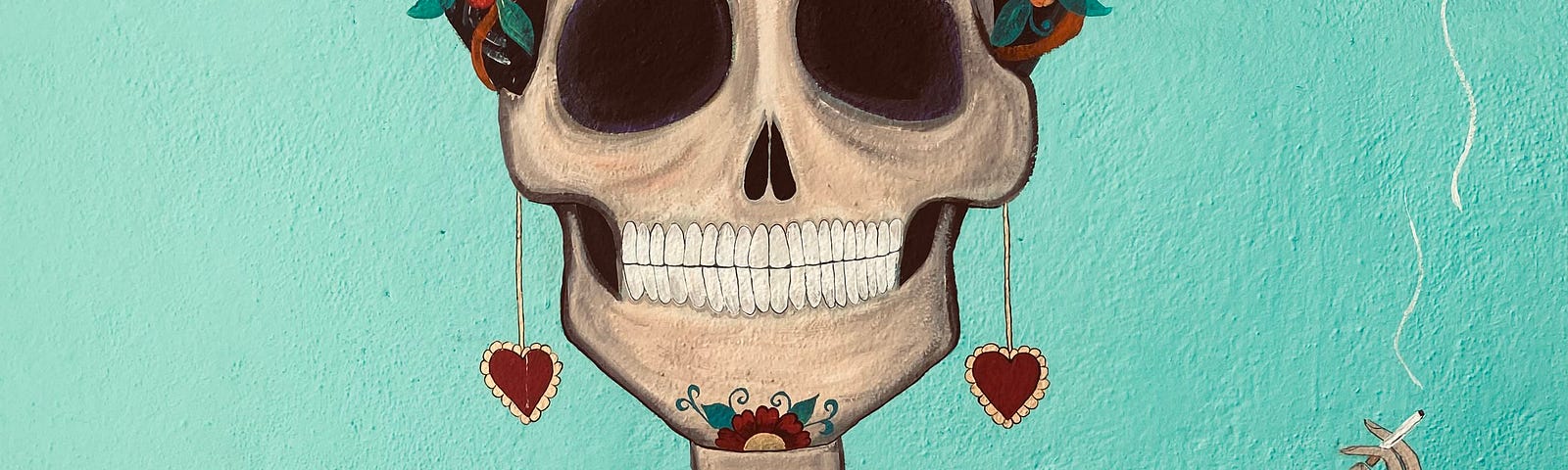 A mural on a blue wall. The mural depicts a sugar skull — a female-appearing skeleton covered in flowers and hearts. She is smoking a cigarette and grinning. Her unibrow suggests that she is Frida Khalo, or at least an homage to her.