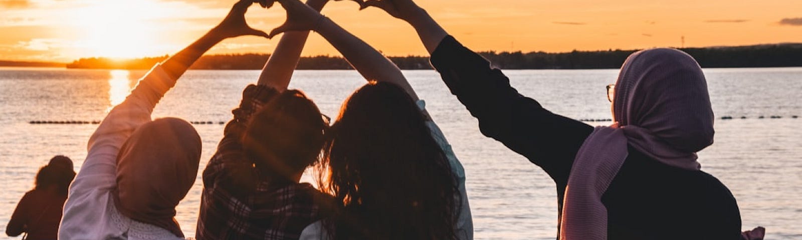 Four friends sitting on the beach at sunset while making the heart signs with their hands.